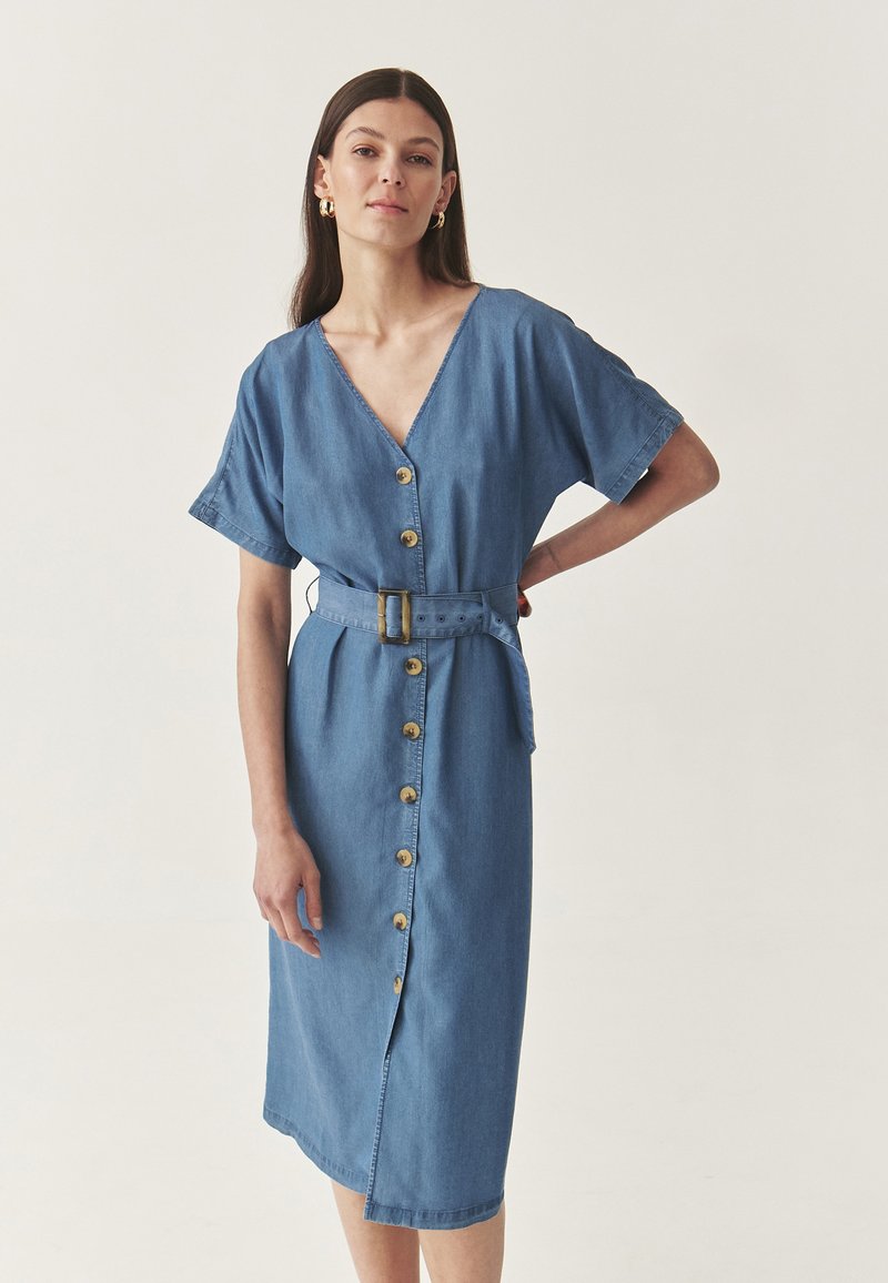 Denimkok dress med korta ärmar, V-ringning, knäppning fram och bälte i midjan. Blått tyg med en slät textur och gyllene knappar.