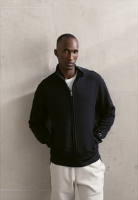 Czarna rozpinana bluza z teksturowanej tkaniny, z wysokim kołnierzem i przednimi kieszeniami, noszona na białej koszuli. Model stoi na neutralnym tle.