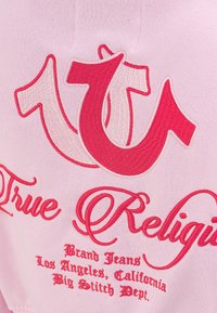 Rosa Stoff mit gesticktem roten und weißen "True Religion"-Logo und Text, einschließlich "Brand Jeans Los Angeles, California Big Stitch Dept."