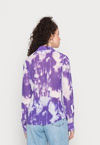 Camisa de manga longa com padrão de tie-dye em roxo, apresentando um colarinho e detalhes de botões, combinada com jeans azul claro, mostrando um ajuste relaxado.