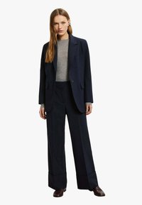 Blazer sartoriale blu navy e pantaloni a gamba larga, abbinati a un maglione grigio. Tessuto liscio con un leggero splendore, con chiusura a un solo bottone.