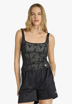 Giovane donna che indossa una canotta nera con motivi e pantaloncini neri larghi con strisce bianche, in piedi con una mano in tasca.