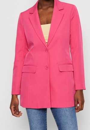 Vrouw draagt een felroze blazer met twee knopen en klepzakken over een beige top en blauwe spijkerbroek, staand tegen een lichte achtergrond.