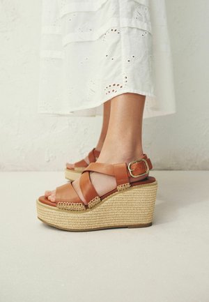 Picioare purtând sandale wedge din piele de culoare tabac cu talpă platformă împletită și catarame aurii, asortate cu o fustă midi albă cu model brodat.