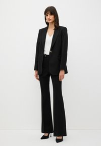 Zwart op maat gemaakte blazer met enkele gouden knoop, gecombineerd met zwarte uitlopers. Witte V-hals top en zwarte puntige hakken maken de look af.