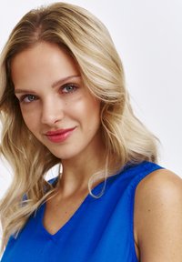 Niebieska bluzka bez rękawów z dekoltem w kształcie litery V. Modelka ma blond, falowane włosy i widoczne makijaż, a jej cera jest gładka.