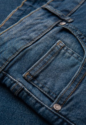 Tessuto di denim blu scuro con tasca, cuciture a contrasto e un piccolo bottone in metallo. La texture appare liscia con un leggero effetto sbiadito.