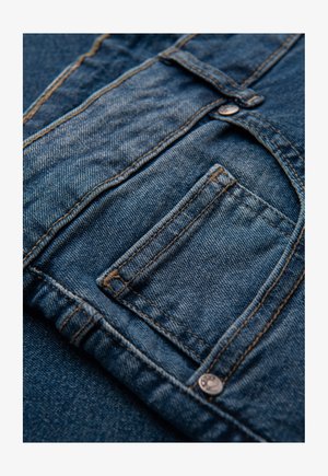 Tessuto di denim blu scuro con tasca, cuciture a contrasto e un piccolo bottone in metallo. La texture appare liscia con un leggero effetto sbiadito.