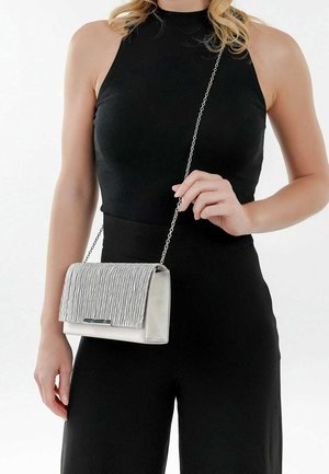 Femme en tenue noire sans manches tenant un sac bandoulière argenté texturé avec une chaîne, sur fond blanc.