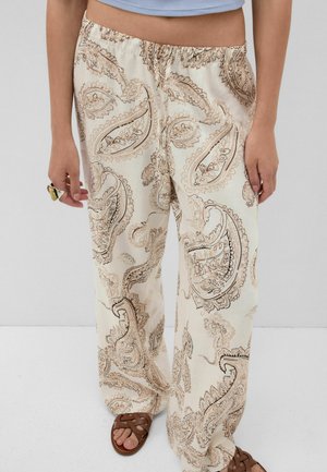 Person iført cremefarvede bukser med vidde og brunt paisleyprint, blå croppet top, brune flettede sandaler og en ring på venstre hånd.