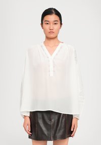 Femme aux cheveux noirs attachés, portant un haut blanc ample avec des détails en dentelle et une mini-jupe en cuir marron foncé, se tenant devant un fond uni.