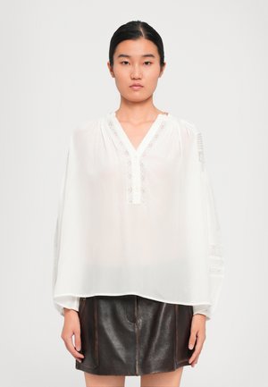 Woman with black hair tied back wearing a loose white blouse with lace details and a dark brown leather mini skirt, standing against a plain background.