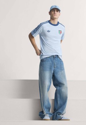 Mand iført lyseblå Adidas-kasket, lyseblå Italiens fodboldtrøje, vidde ben falmede jeans med striber og hvide sneakers, der står med hænderne bag ryggen.