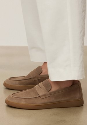 Lysebrune mokkasiner i ruskind med gummisåler, båret med hvide bukser, vist fra siden på en glat beige gulv.