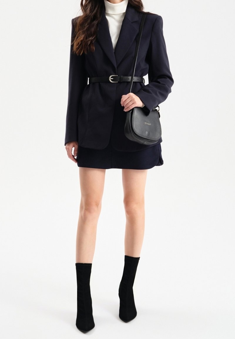 Blazer bleu marine avec une coupe ajustée et une ceinture, col roulé blanc, jupe bleue marine, bottines en suède noir à la cheville, et un petit sac bandoulière noir.