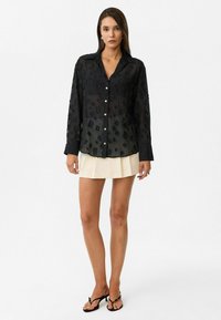 Vrouw in een doorschijnende zwarte blouse met lange mouwen en knopen, met vierkante patronen, een crèmekleurige geplooide minirok en zwarte hoge hakken.