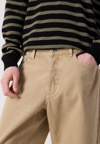 Pantalon beige avec une coupe décontractée, doté d'une fermeture éclair et d'un bouton. Le tissu a une texture lisse, avec des détails de couture visibles.