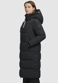 Zwarte pufferjas met een lange stijl, een capuchon, elastische manchetten, ritszakken en een logo-accent aan de voorkant. Glad, gewatteerd textuur.