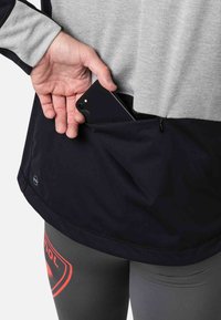 Schwarzes Shirt mit einer Reißverschlusstasche für ein Smartphone, kombiniert mit grauen Leggings mit einem roten Logo. Weicher Stoff, lässiges Design.