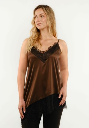 Žena s dlhými blond vlasmi, ktorá má na sebe hnedý saténový top s čiernou čipkovou lemovkou a čierne nohavice, pozrie sa stranou.