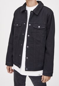 Veste en denim noire avec un col en polaire noire texturée, poches avant et boutons en métal, portée sur une chemise blanche et un pantalon noir.