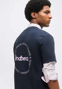 Άνδρας με μπλε σκούρο μπλουζάκι με ροζ κυκλικό κείμενο και γυρισμένα λευκά μανίκια που κοιτάει πλάγια σε μονόχρωμο φόντο.