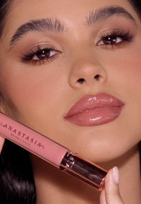 Flüssiger Lippenstift in einer pinken Tube mit goldenen Akzenten. Das Model zeigt glänzende nude Lippen und neutrales Make-up, mit definierten Augenbrauen und Wimpern.