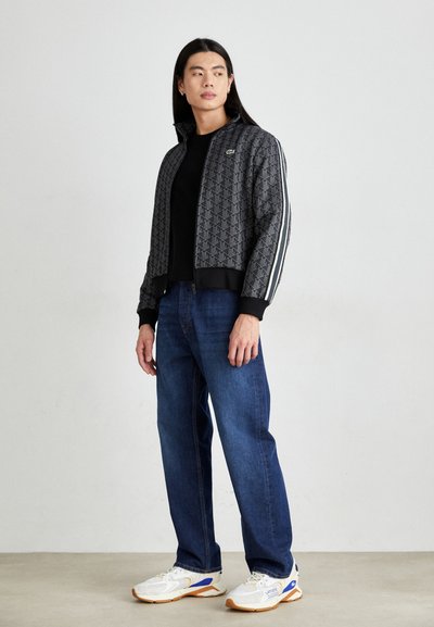 Veste bomber noire avec un motif géométrique, des poignets côtelés et des bandes blanches sur les côtés, associée à un jean bleu et des baskets blanches avec des accents bleus.