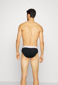 Schwarze Baumwollslips mit einem weißen, gebrandeten elastischen Bund, die eine glatte Textur und eine figurbetonte Passform aufweisen, von hinten betrachtet.