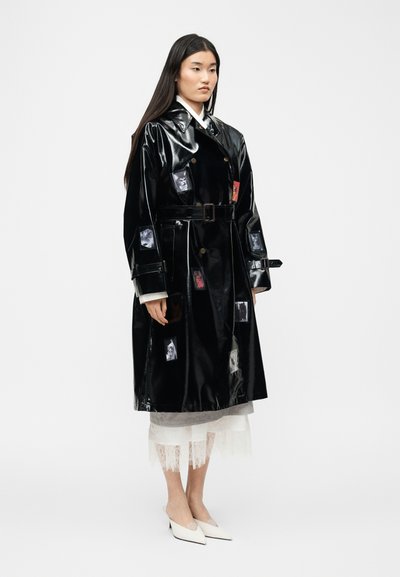 Γυναίκα στέκεται φορώντας μία γυαλιστερή μαύρη καμπαρντίνα με ζώνη και φωτογραφικά patches, μια λευκή δαντελένια φούστα και λευκά μυτερά παπούτσια σε απλό φόντο.