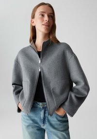 Jonge vrouw in een grijze wollen zip-up Jack met zakken en blauwe spijkerbroek, staand met haar handen in de zakken, kijkend met een lichte opwaartse blik.