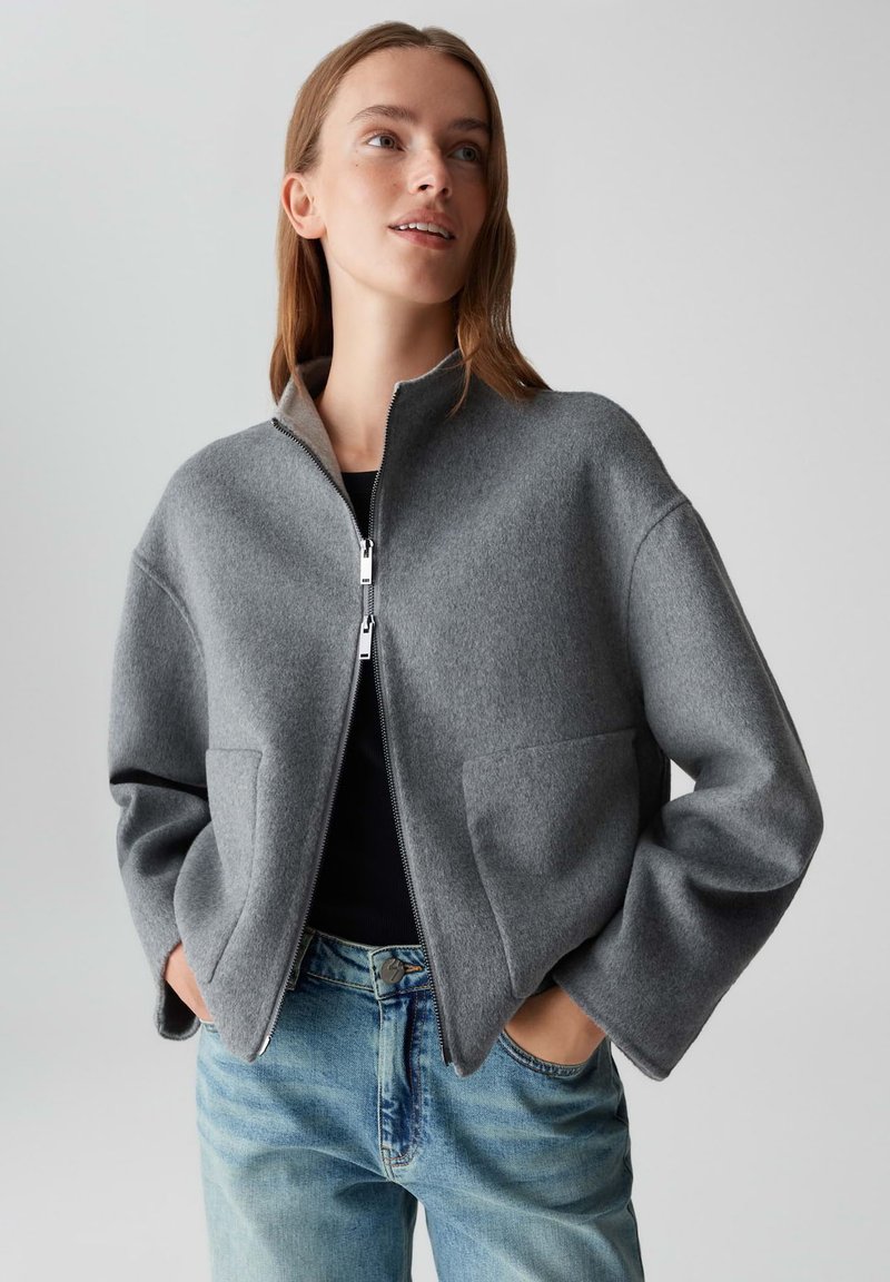 Jonge vrouw in een grijze wollen zip-up Jack met zakken en blauwe spijkerbroek, staand met haar handen in de zakken, kijkend met een lichte opwaartse blik.
