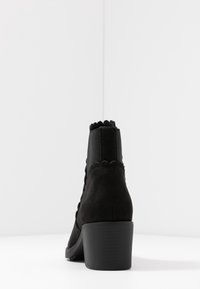 Czarne zamszowe botki do kostek z falbaniastym brzegiem, elastycznymi panelami bocznymi i średnim obcasem w kształcie bloku. Gładka faktura i minimalistyczny design.