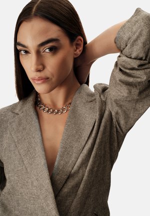 Blazer grigio testurizzato con scollo a V profondo e maniche raggruppate, abbinato a una collana a catena d'argento. Tonalità neutre e design elegante.