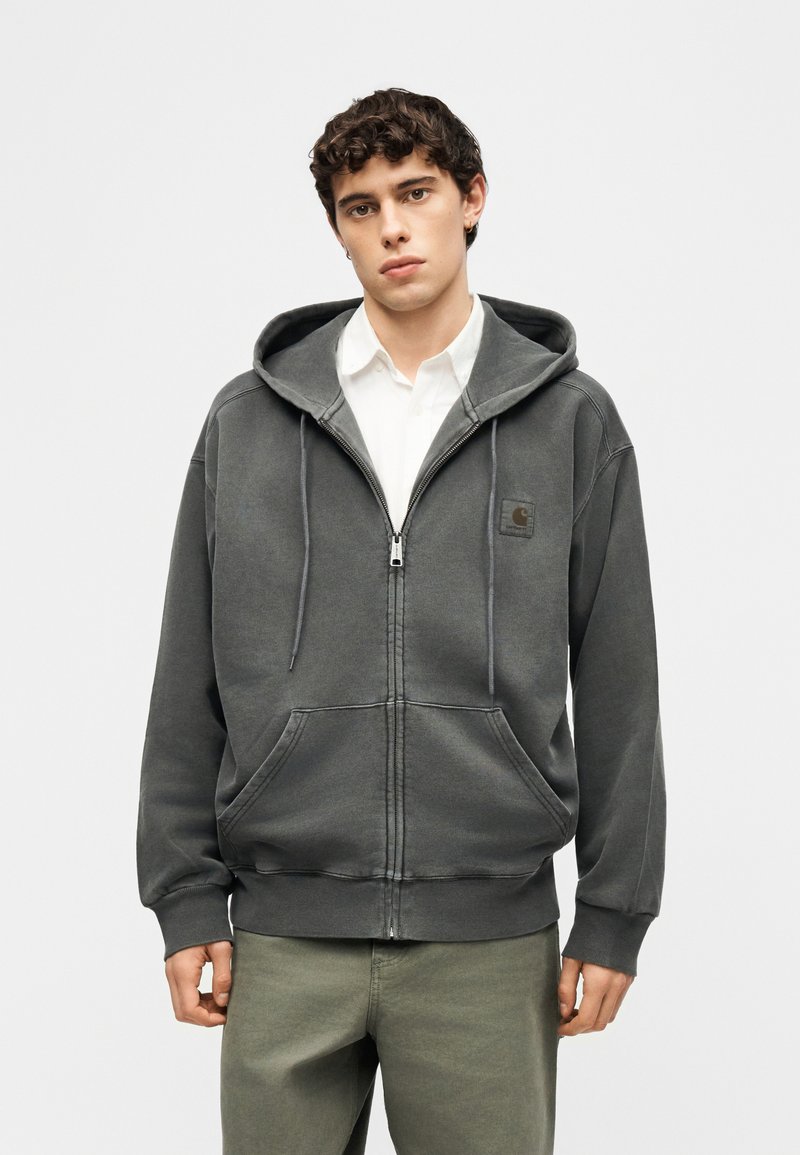 Young man with short curly hair wearing a dark gray zip-up hoodie over a white shirt and olive green pants, standing against a plain background.