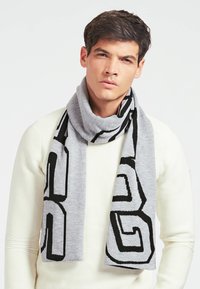 Écharpe en mélange de laine grise avec un audacieux motif géométrique noir. L'écharpe est enroulée autour du cou pour un look superposé.