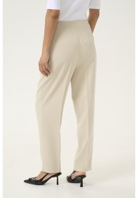 Pantalon taille haute beige avec une coupe ajustée, en tissu lisse et avec des détails sur les coutures latérales, associé à des escarpins noirs à bouts pointus.