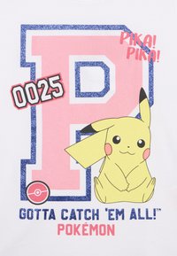 Balsam baltā grafiskā T-kreklā ar lielu rozā burtu "P", kas ietverts zilā spīdīgā kontūrā, Pikachu ilustrācija un teksts "Jānoķer visi!"