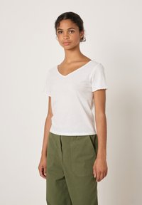 Valge lühikeste varrukatega V-kaelusega T-särk, valmistatud pehmest kangast, kombineeritud oliivroheliste, lõdva lõikega pükstega, millel on plaastritaskud ja vertikaalsed triibud.