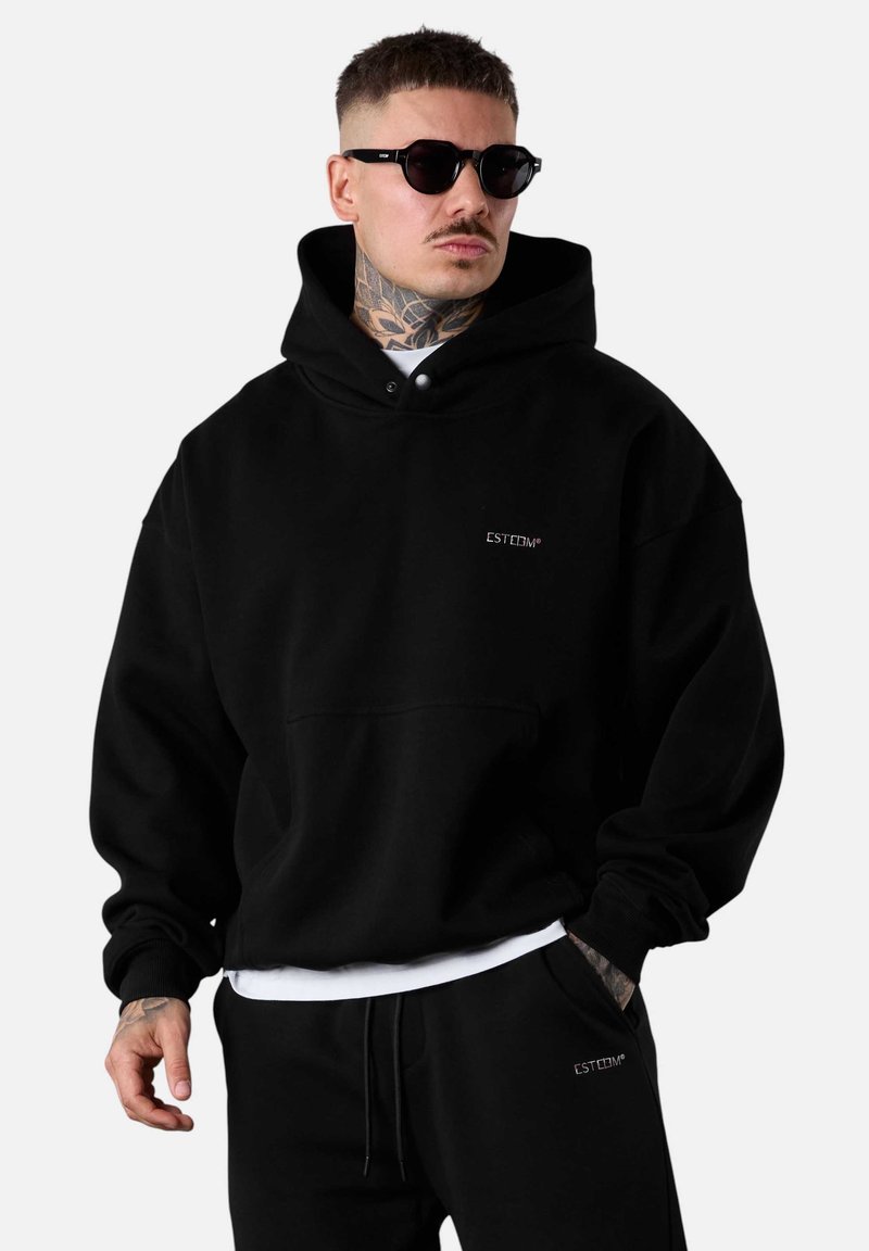 Sweatshirt à capuche noire avec une poche avant et un logo brodé. Le mannequin porte des lunettes de soleil et un jogging foncé. Texture lisse.