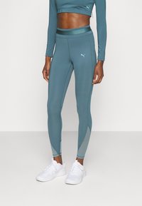 Kvinna i teal höga leggings med nätpaneler och en kortärmad topp, som matchar med vita sportskor.