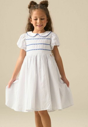 Jeune fille aux cheveux bouclés coiffés en chignons, portant une robe blanche avec un bord bleu et de petits pois, tenant les côtés de la robe et souriant doucement.
