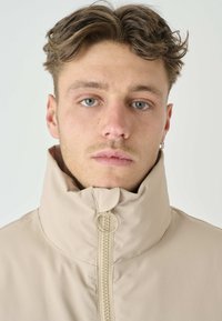 Beige leichte Jacke mit hohem Kragen, Frontreißverschluss und Ringgriff. Glatte Textur mit minimal sichtbaren Nähten.