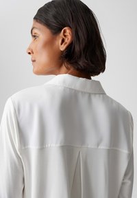 Weiße Bluse aus einem weichen Stoff mit umschlagbarem Kragen und einer Rückenfalte. Die Bluse wird von der Seite gezeigt, wodurch der Ausschnitt und die Schulter betont werden.