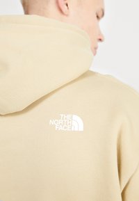 Sudadera con capucha beige con una textura suave, bolsillo tipo canguro y un logo blanco en la parte trasera. Diseño simple sin patrones.