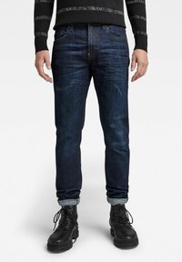 Jeans de mezclilla azul oscuro con un corte ajustado, que presentan sutiles patrones de desgaste, dobladillos remangados y una textura general suave. Llevados con botas negras.