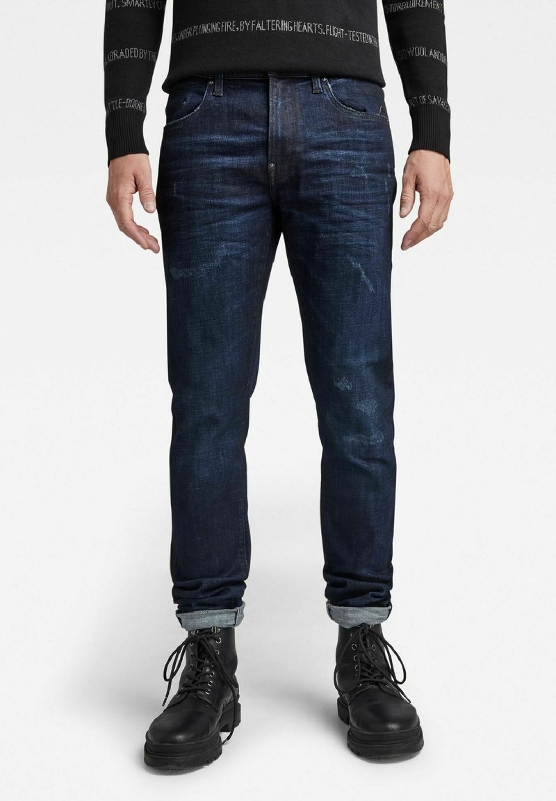 Jeans de mezclilla azul oscuro con un corte ajustado, que presentan sutiles patrones de desgaste, dobladillos remangados y una textura general suave. Llevados con botas negras.