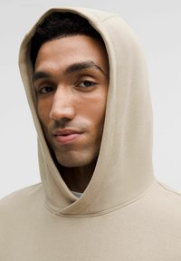 Beige huppari, jossa on suuri huppu, valmistettu teksturoidusta kankaasta. Ominaisuuksiin kuuluu näkyvät saumat ja rento malli, alla on yksinkertainen valkoinen paita.