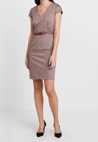 Robe marron à pois avec manches courtes, encolure en V, coupe ajustée et ceinture marron à la taille. Portée avec des chaussures à talons hauts noires.