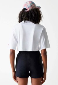Witte cropped t-shirt met korte mouwen, gecombineerd met zwarte elastische shorts. Een roze en blauwe geruite pet wordt gedragen. Zachte stofstructuren zijn zichtbaar.